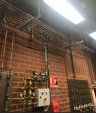 Photo of a red brick wall covered with piping with gauges and a control panel with buttons.