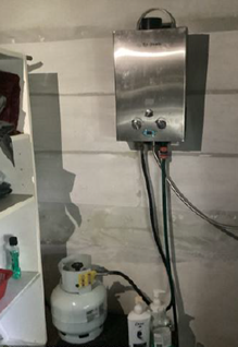 Photo showing a silver gas water heater against a wall connected to a white LP gas cylinder bottle on the floor beneath it.